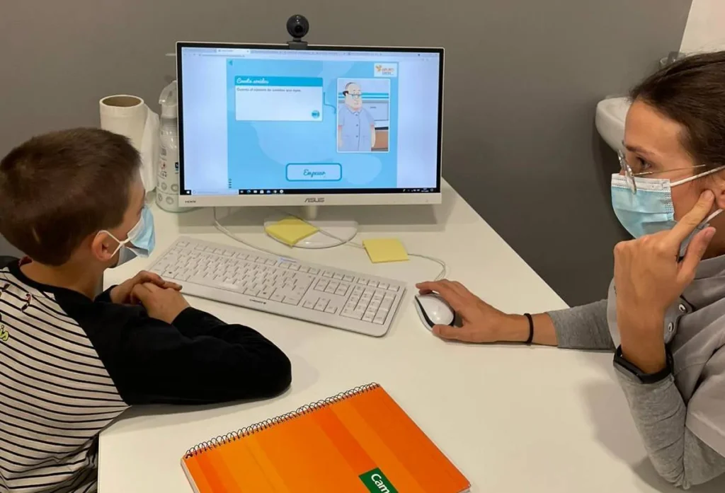 Criança em frente a um computador conta estímulos sonoros; adulto ao lado explica, ambos com máscaras, ambiente educativo