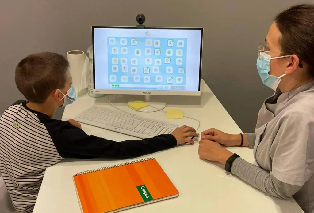 Criança em frente a um computador com ícones na tela, supervisionada por uma mulher com máscara; caderno laranja e rolo de papel na mesa.