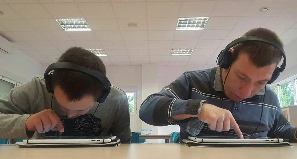 Duas pessoas com deficiência intelectual usam fones de ouvido e tablets, interagindo com dispositivos digitais em uma sala de aula.