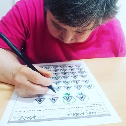 Pessoa com deficiência intelectual escreve com caneta preta em uma folha com símbolos; camiseta rosa e mesa.