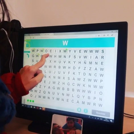 Tela de computador com jogo de palavras em grade; uma pessoa aponta a letra 'W' e há uma videochamada com duas pessoas na parte inferior.