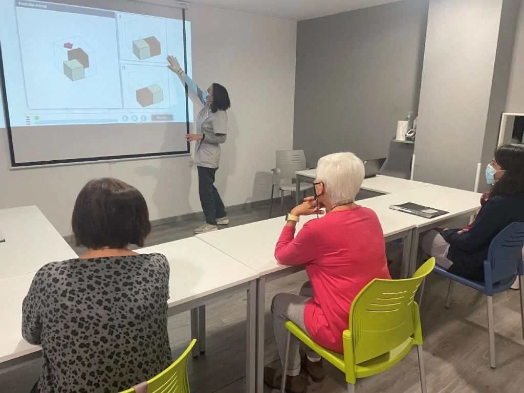 Quatro participantes observam uma pessoa em frente a uma tela, apontando; cubos sobre a mesa em uma sala de aula com cadeiras coloridas e máscaras.