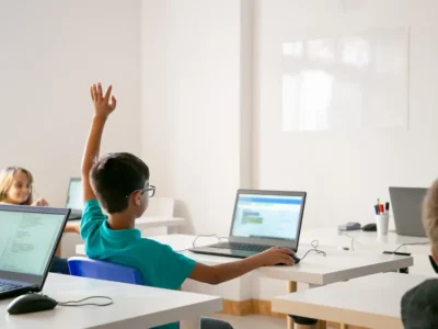 Menino de origem mestiça com óculos levanta a mão em uma sala de aula luminosa, cercado por colegas e laptops