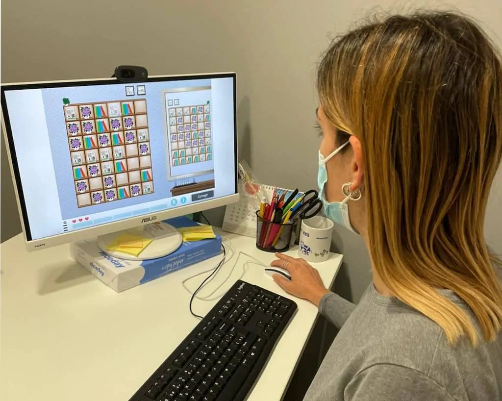 Profissional orienta uma pessoa em frente a um computador com máscara; a tela mostra uma tarefa visual com grades de imagens.