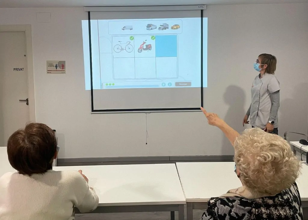 Mulher de pé em frente a uma tela com a atividade Ordenar sequência do NeuronUP; duas pessoas observam.