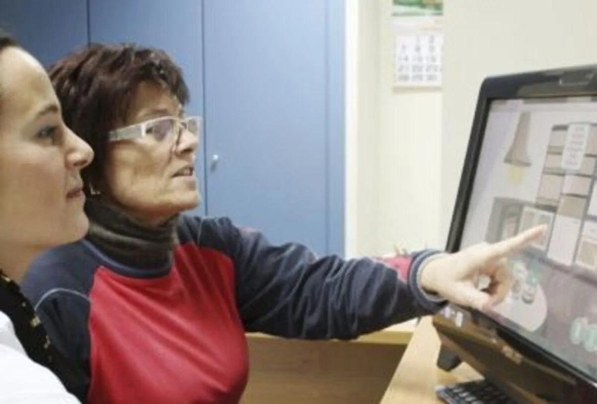 Duas mulheres frente a um computador em escritório, uma aponta para a tela enquanto explica; ambiente com armários ao fundo.