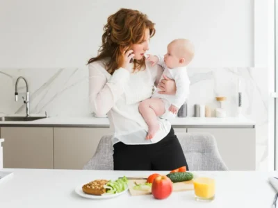 Mulher de cabelo cacheado segura um bebê enquanto fala ao telefone em uma cozinha moderna, exercitando a atenção alternante.