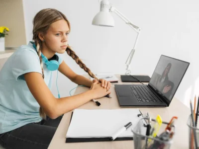 Jovem com tranças usando fones de ouvido azuis diante de um laptop em uma escrivaninha bem iluminada, com uma lâmpada, caderno e planta.
