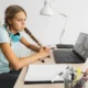 Jovem com tranças usando fones de ouvido azuis diante de um laptop em uma escrivaninha bem iluminada, com uma lâmpada, caderno e planta.