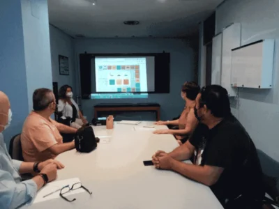 Reunião de equipe em sala de conferência: cinco pessoas ao redor da mesa olhando para uma tela com conteúdo digital em um ambiente clínico.