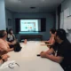 Reunião de equipe em sala de conferência: cinco pessoas ao redor da mesa olhando para uma tela com conteúdo digital em um ambiente clínico.