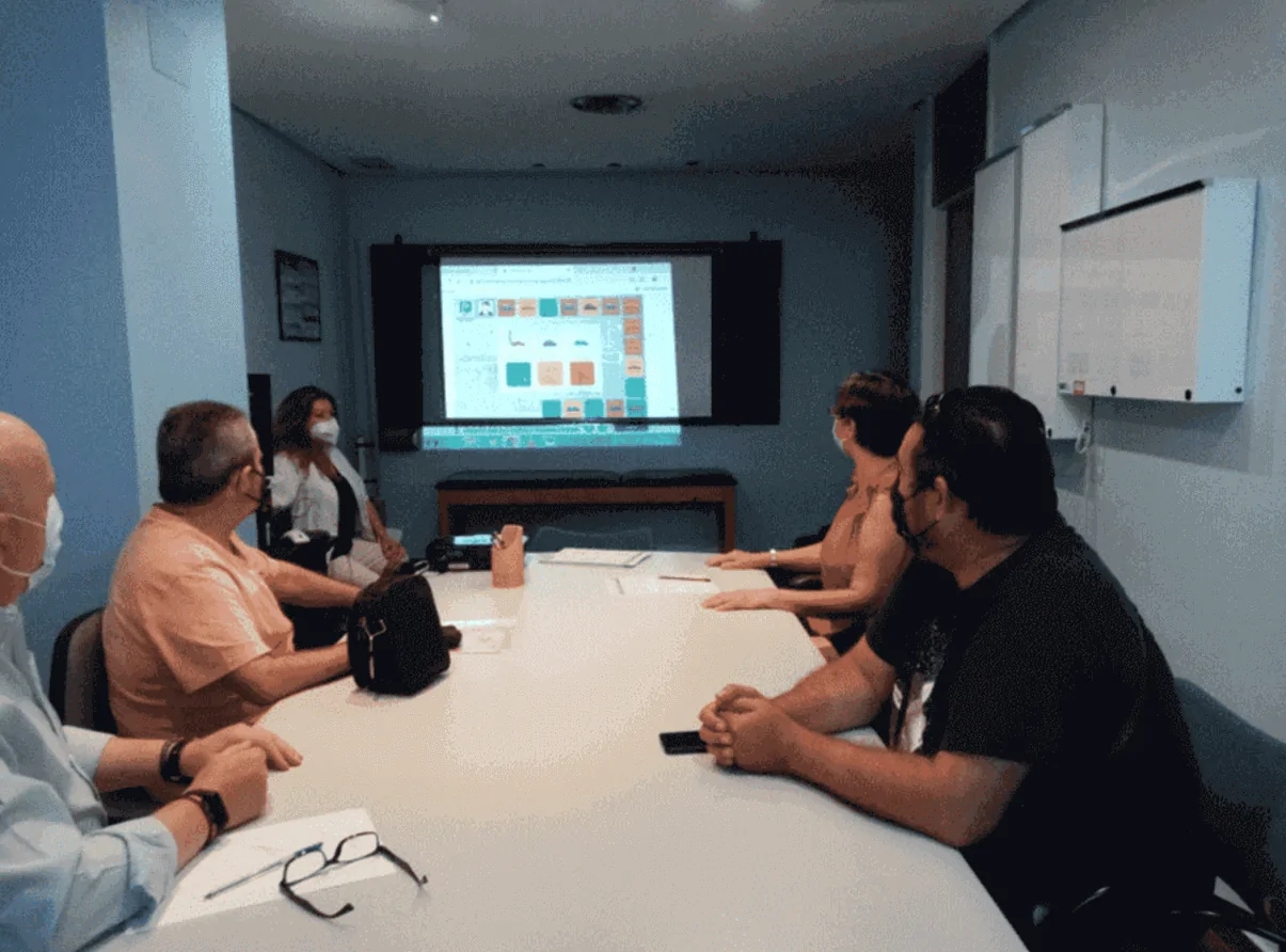 Reunião de equipe em sala de conferência: cinco pessoas ao redor da mesa olhando para uma tela com conteúdo digital em um ambiente clínico.