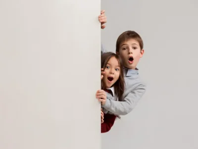 Duas crianças espiando por trás de uma parede branca, com expressões de surpresa, em um banner educativo visível.