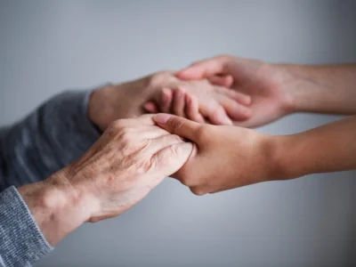 Duas mãos entrelaçadas, uma idosa com rugas e a outra mais jovem, sobre fundo neutro.