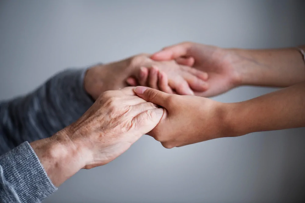 Duas mãos entrelaçadas, uma de idade mais avançada com rugas e a outra mais jovem, sobre fundo neutro.
