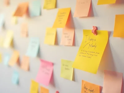 Parede coberta de notas adesivas coloridas (amarelo, rosa e laranja) para organizar ideias e tarefas, com uma nota em destaque que parece dizer 'Tilly's Menu'.