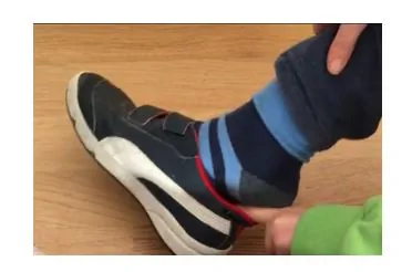 Niño pequeño sentado de perfil ajustando una zapatilla oscura con Velcro; calcetas azules y pantalón azul, en casa.