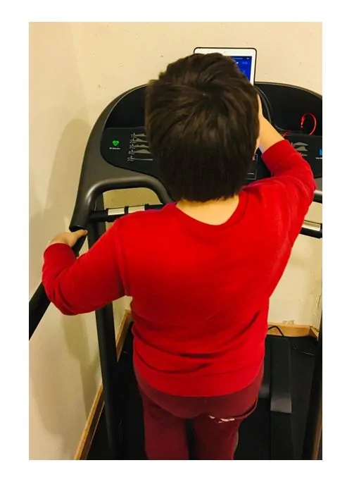 Niño visto desde atrás sobre una caminadora en un entorno de rehabilitación motora infantil, mirando una pantalla durante un ejercicio.