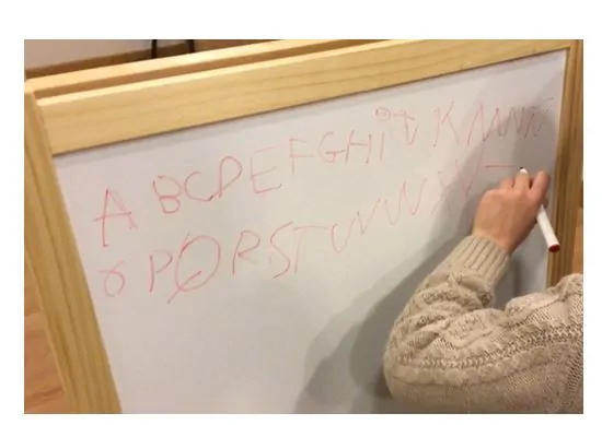 Mujer trabajando lectoescritura frente a pizarra, con letras en rojo y mano escribiendo, en un aula para ejercicios de intervención.
