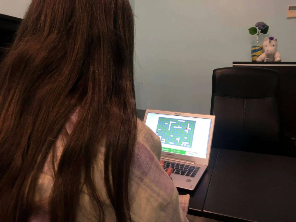 Pessoa com cabelo longo sentada em frente a um portátil, joga um labirinto na tela; fundo verde, planta e pelúcia.