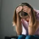 Plano fechado de uma jovem com expressão triste, segurando a cabeça com as duas mãos diante de um fundo neutro.