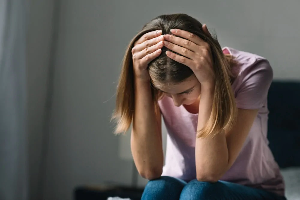 Primeiro plano de uma jovem com expressão triste, segurando a cabeça com as duas mãos diante de um fundo neutro.