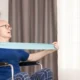 Idoso sentado em uma cadeira de rodas realiza exercícios com faixa de resistência azul em casa, supervisionado por uma enfermeira.