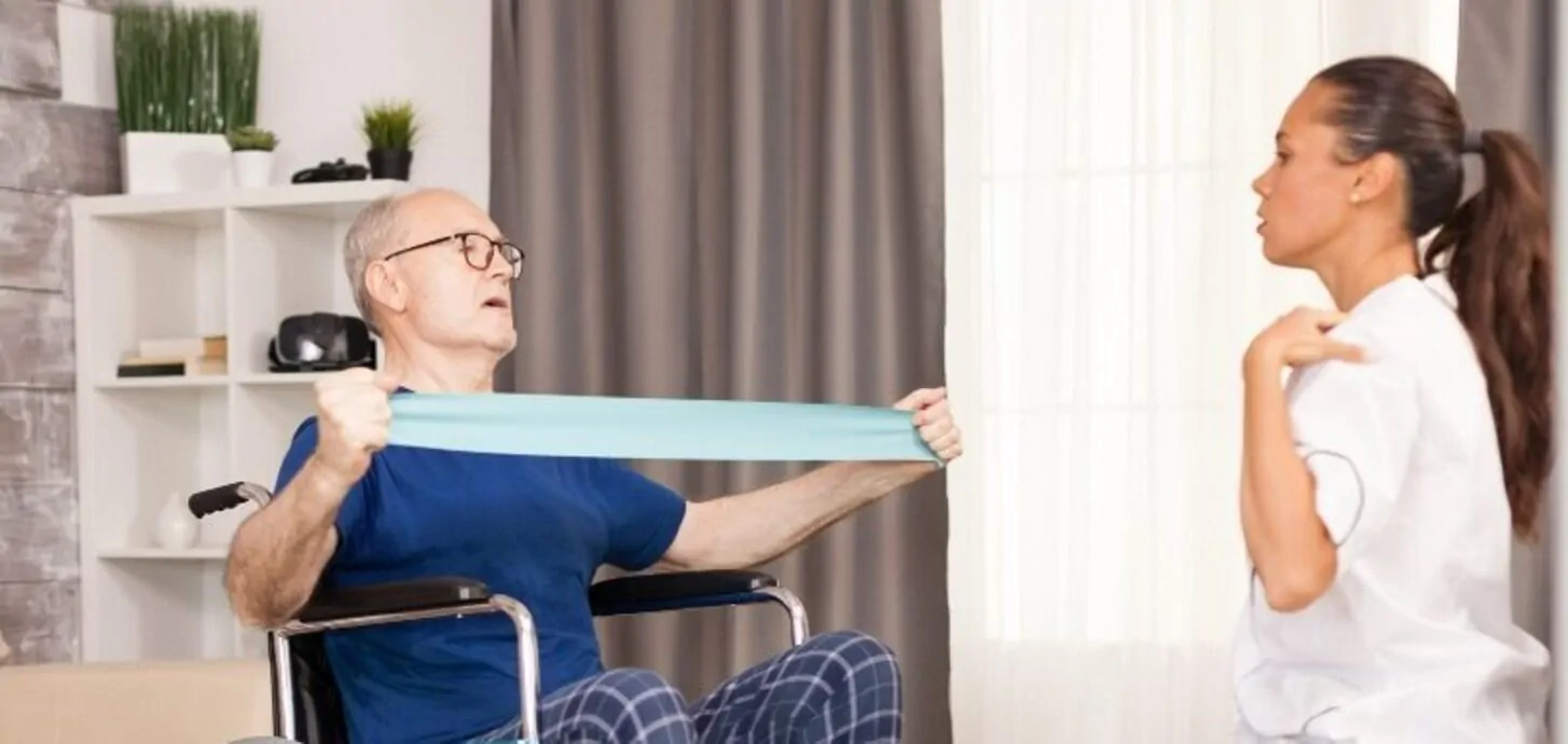 Idoso sentado em uma cadeira de rodas realiza exercícios com faixa de resistência azul em casa, supervisionado por uma enfermeira.