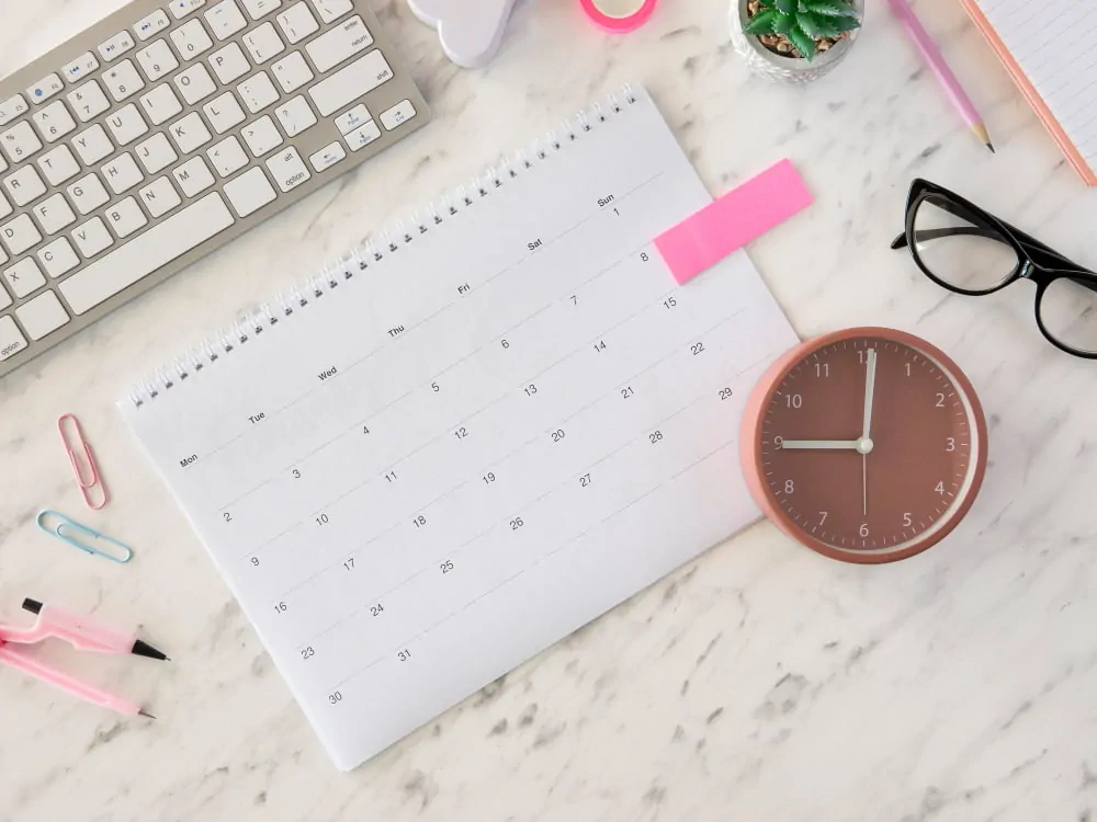 Plano cenital de uma mesa de trabalho organizada com calendário, relógio rosa, laptop, óculos e planta, com foco na gestão do tempo.