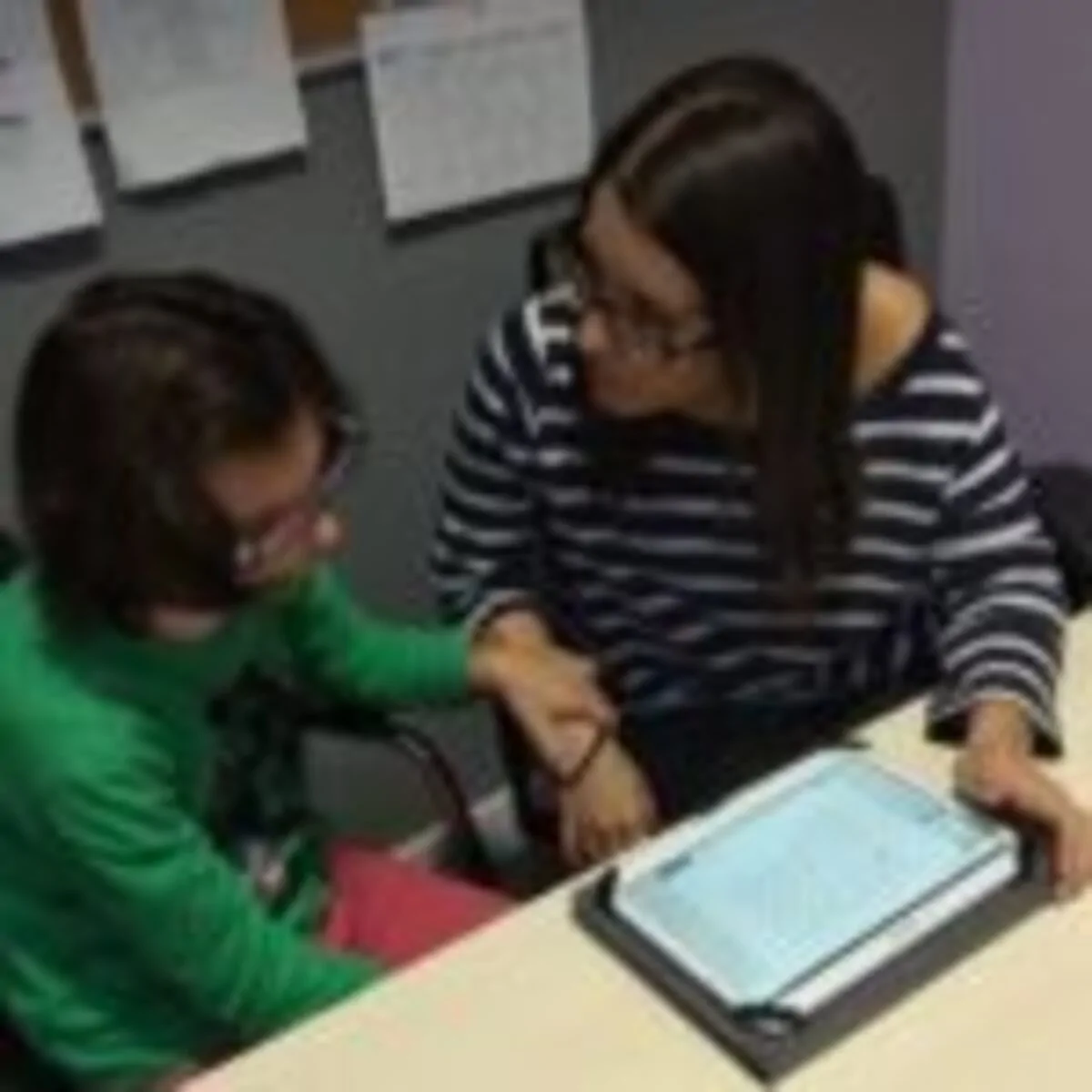 Mulher e menina sentadas à mesa olhando para um tablet, em um ambiente educacional, com apoio e interação visíveis, relacionado com a síndrome 5p-.
