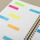 Caderno aberto visto de cima com páginas em branco e marcadores coloridos; espiral dourada lateral e fundo neutro.