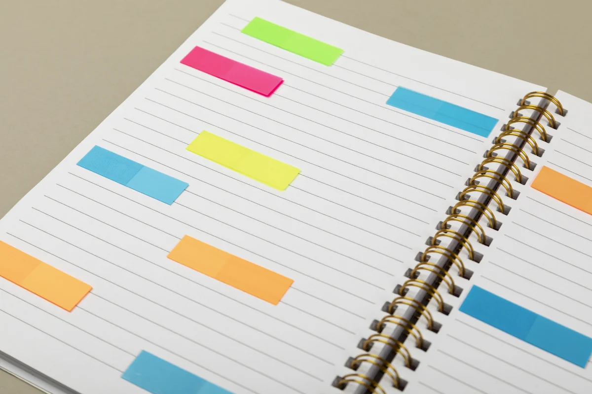 Caderno aberto visto de cima com páginas em branco e marcadores coloridos; espiral dourado lateral e fundo neutro.