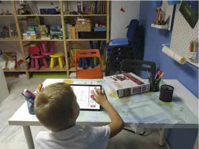 Menino sentado diante de um tablet e de uma folha sobre uma mesa branca, dentro de uma sala de aula organizada para exercícios de estimulação neuropsicológica da dislexia.
