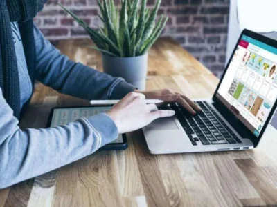 Profissional de neuropsicologia trabalhando à distância: notebook, tablet e stylus, com uma planta e uma parede de tijolos ao fundo.