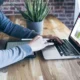 Profissional de neuropsicologia trabalhando à distância: notebook, tablet e stylus, com uma planta e uma parede de tijolos ao fundo.