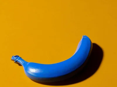 Banana azul pintada sobre um fundo amarelo, criando um contraste visual chamativo e um toque surrealista na diapositiva.