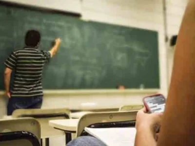Sala de aula com professor de costas para o quadro verde; primeiro plano de aluno sentado com telefone celular, ambiente de sala de aula.