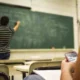 Sala de aula com professor de costas para o quadro verde; primeiro plano de aluno sentado com telefone celular, ambiente de sala de aula.