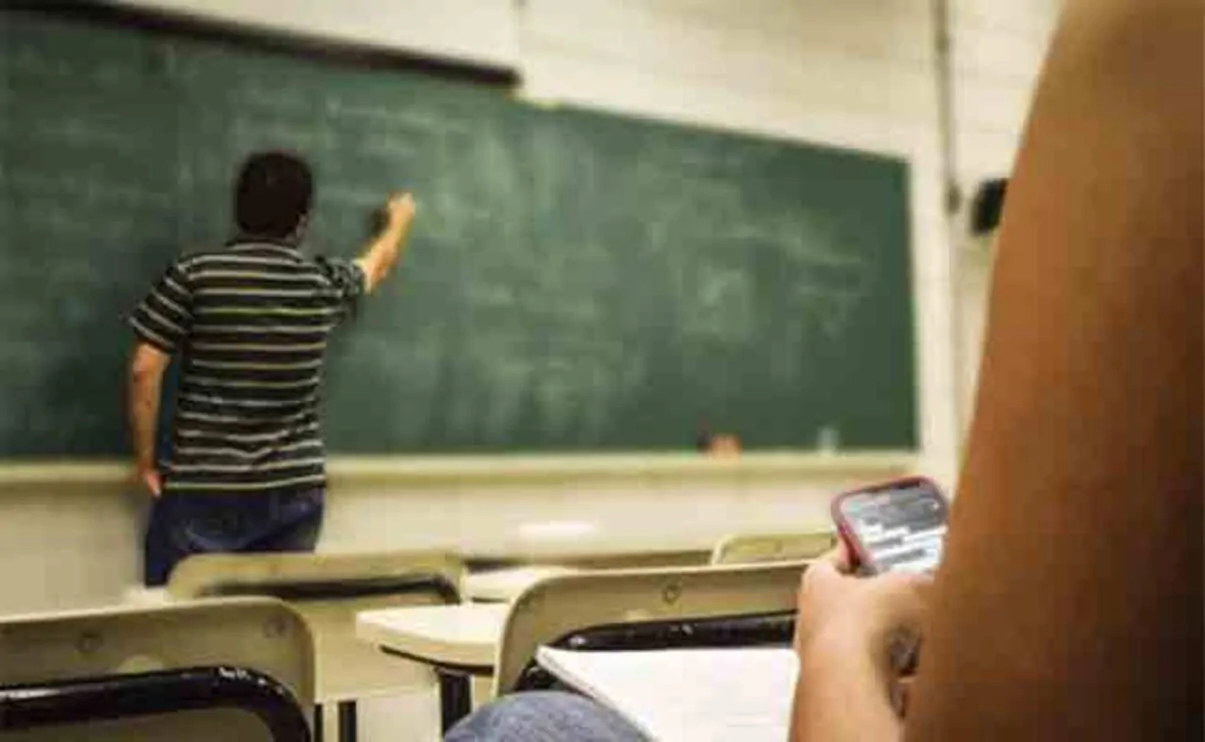 Sala de aula com professor de costas para um quadro verde; primeiro plano de estudante sentado com telefone celular, ambiente de sala de aula.