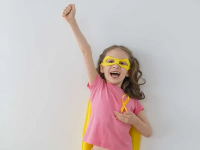 Menina vestida de superheroína com o laço dourado do Dia Mundial da Criança com Câncer, olhando para a câmera com expressão esperançosa.