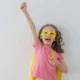 Menina vestida de superheroína com o laço dourado do Dia Mundial da Criança com Câncer, olhando para a câmera com expressão esperançosa.