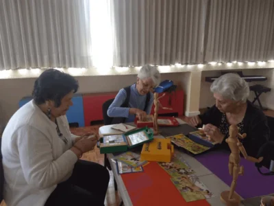 Grupo de três mulheres idosas sentadas ao redor de uma mesa, realizando atividades com materiais em um ambiente luminoso e acolhedor.