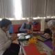 Grupo de três mulheres idosas sentadas ao redor de uma mesa, realizando atividades com materiais em um ambiente luminoso e acolhedor.