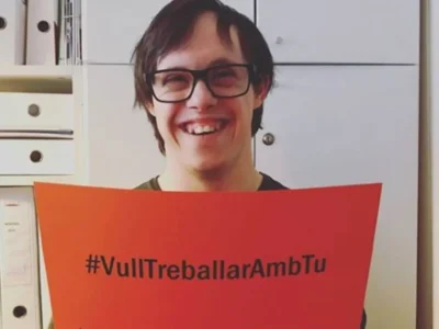 Pessoa sorridente com óculos segura cartaz laranja com '#VullTreballarAmbTu' e '#QuieroTrabajarContigo' em um escritório.