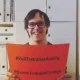 Pessoa sorridente com óculos segura cartaz laranja com '#VullTreballarAmbTu' e '#QuieroTrabajarContigo' em um escritório.