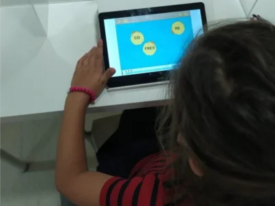 Menina diante de um tablet com fundo azul e palavras em círculos, mostrando exercícios de fonoaudiologia em um ambiente educacional.