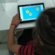 Menina diante de um tablet com fundo azul e palavras em círculos, mostrando exercícios de fonoaudiologia em um ambiente educacional.