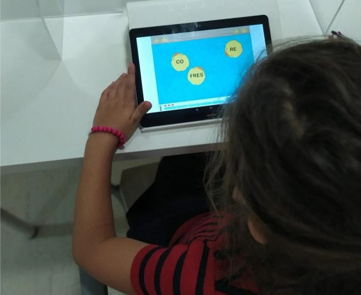 Menina em frente a um tablet com fundo azul e palavras em círculos, mostrando exercícios de fonoaudiologia em um ambiente educativo.