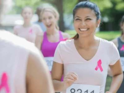 Grupo de mulheres correndo em uma corrida ao ar livre, com camisetas cor-de-rosa e dorsais, em um ambiente verde.