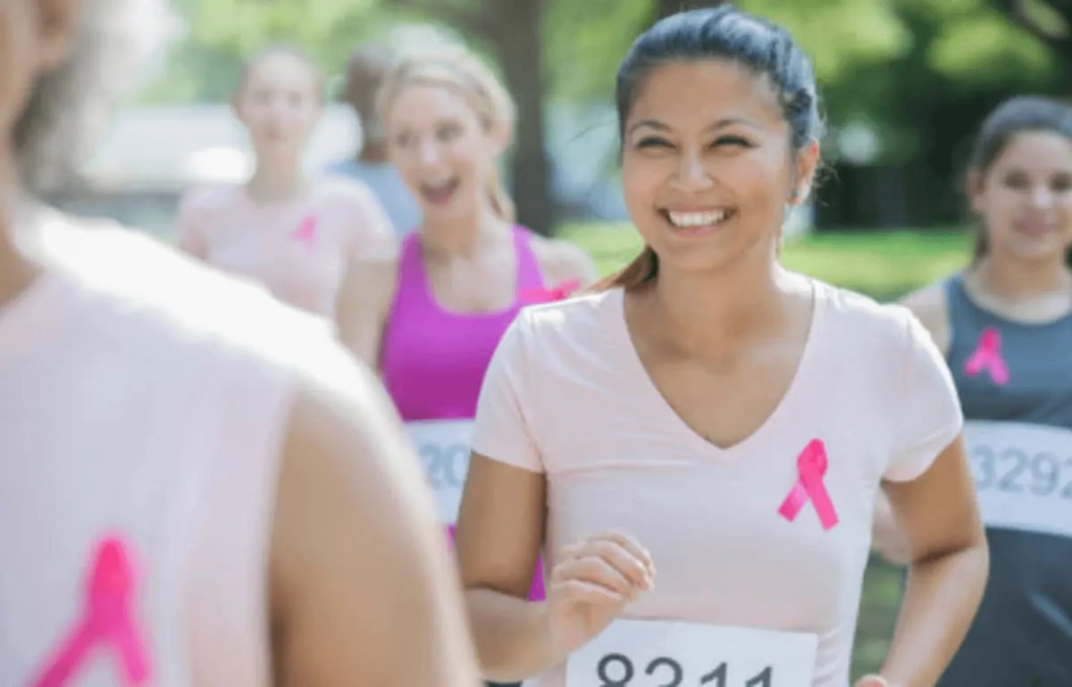 Grupo de mulheres correndo em uma corrida ao ar livre, com camisetas rosas e dorsais, em um ambiente verde.